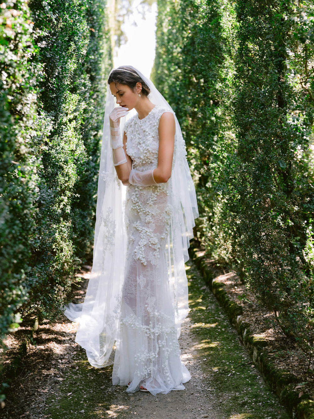 Woman wearing Monique Lhuillier Fall 2024 white fitted sleeveless 3-d embroidered Gabrielle gown with matching veil and tulle gloves