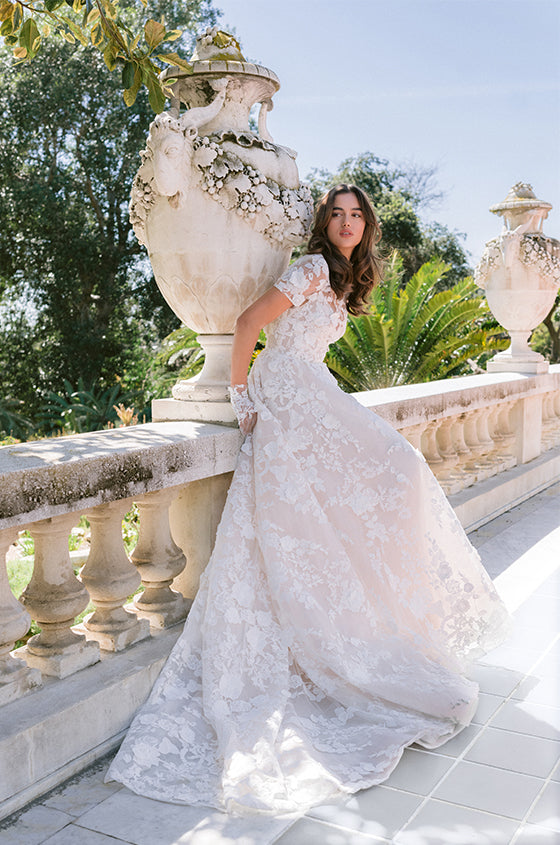 Woman wearing Monique Lhuillier Spring 2024 blush embroidered a-line Antoinette gown with short sleeve embroidered jacket and lace gloves