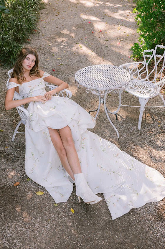 Woman wearing Monique Lhuillier Spring 2024 off-the-shoulder draped neckline high-low white and floral printed Soleil ballgown