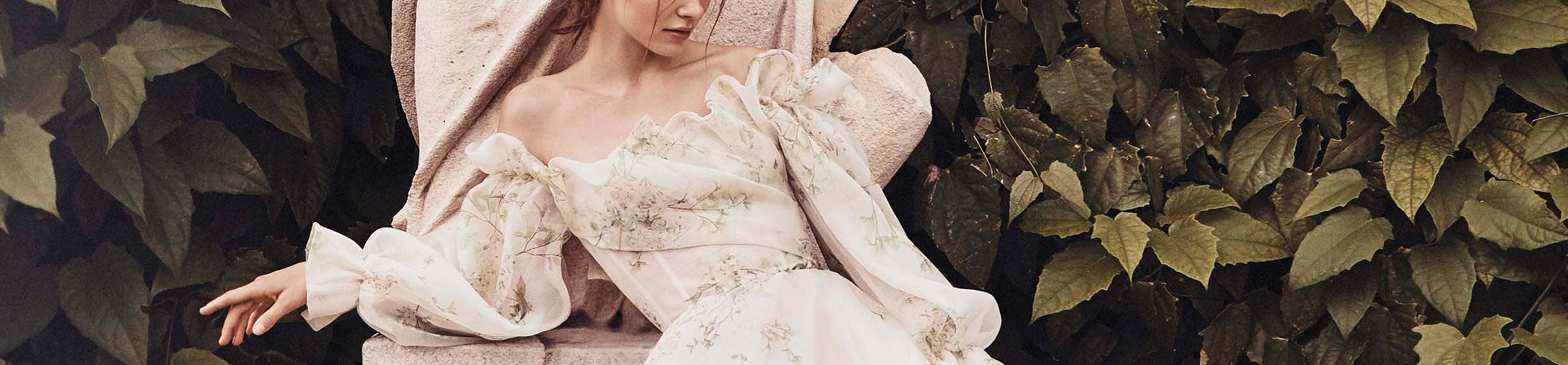 Model leaning on statue wearing ivory and green floral printed off-the-shoulder gazar Bloom a-line gown with ruffle neckline and billowed sleeve 