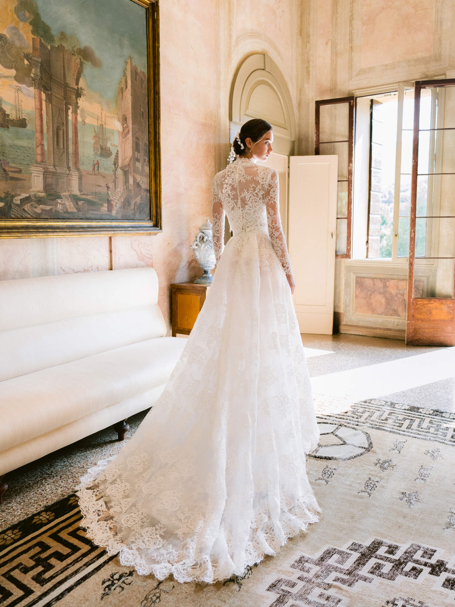 Woman wearing Monique Lhuillier Fall 2024 white Melrose lace bralette with lace long-sleeve high neck jacket and lace a-line skirt with slit