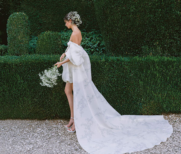 Monique Lhuillier Fall 2024 strapless white and leaf printed gazar bubble hem Avery
cocktail gown with detachable draped sleeve cape train on model in front of greenery