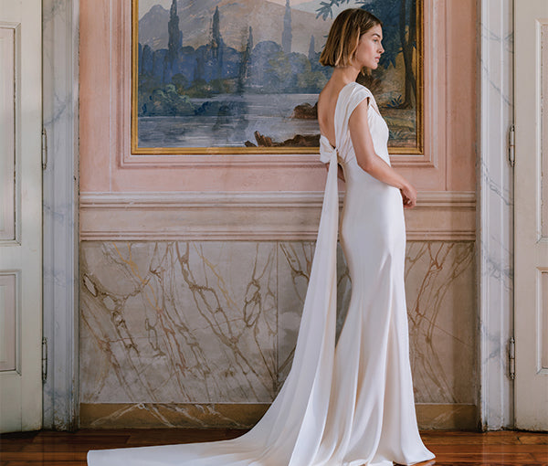 Monique Lhuillier Fall 2025 ivory crepe Rowe sheath gown with asymmetrical neckline on model in front of painting
