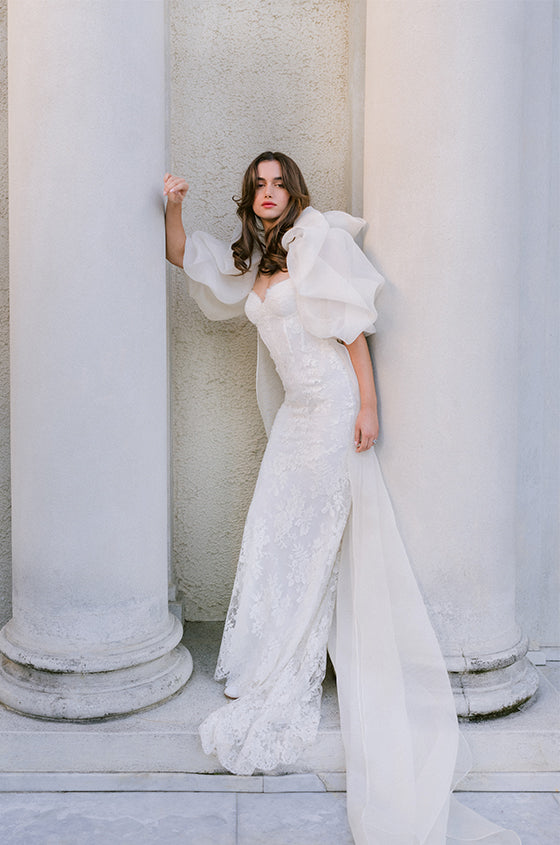 Woman wearing Monique Lhuillier Spring 2024 strapless white lace corseted Eden slip gown with puffed sleeve Barrington coat