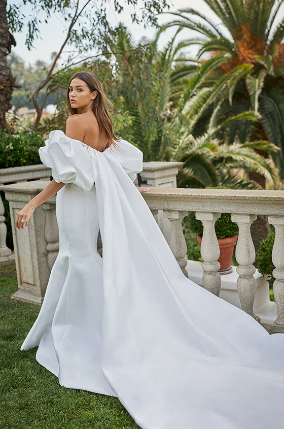 Woman wearing white Monique Lhuillier Spring 2021 mikado fitted off-the-shoulder Admire gown with Admire cape