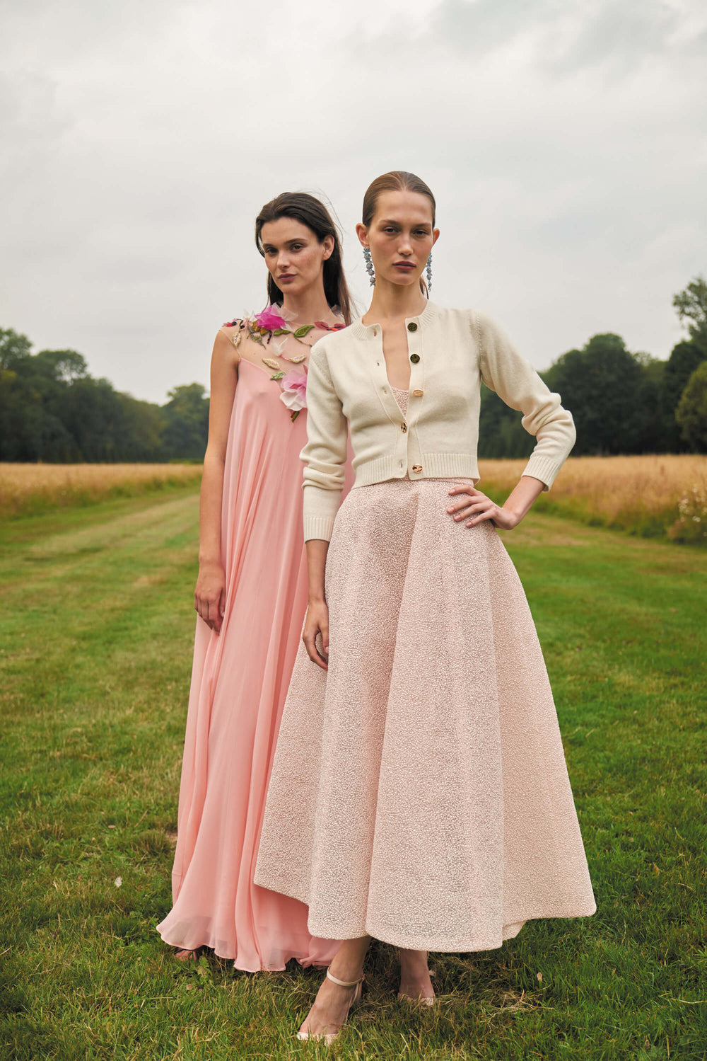 Monique Lhuillier Spring 2024 Melon chiffon caftan gown with floral 3-D embroidery over an illusion tulle neckline next to a powder pink strapless cocktail dress in a field.