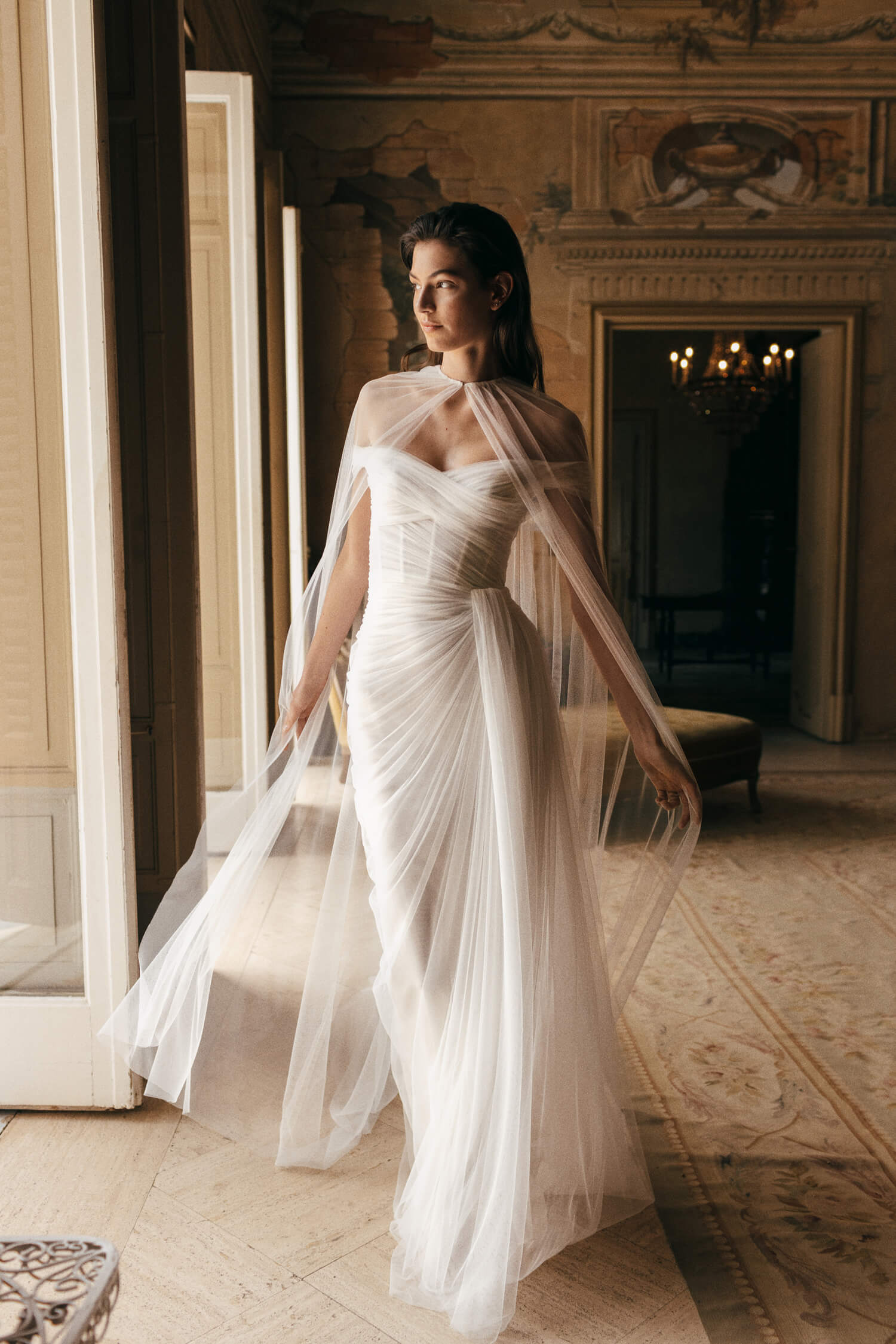 Woman wearing white Spanish tulle draped off-the-shoulder column Monique Lhuillier Spring 2023 Sienna gown with matching Sienna cape