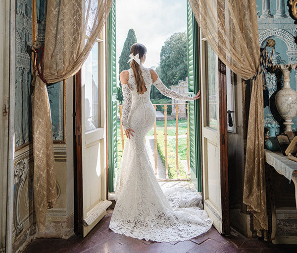 Monique Lhuillier Spring 2025 ivory lace halter Hayworth trumpet gown with lace Hayworth gauntlet sleeves on model looking out window