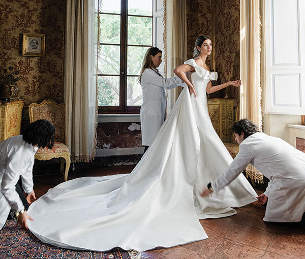 Monique Lhuillier Spring 2025 white mikado off-the-shoulder Mayfair trumpet gown with bow detail at neckline and train on model by windo