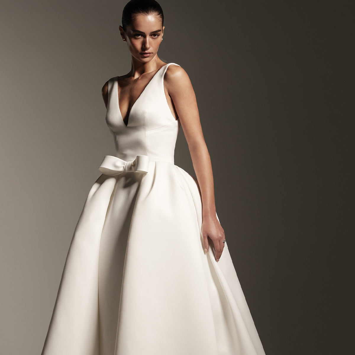 Model wearing Monique Lhuillier Spring 2026 Bridal white matte satin v-neck Avalon column gown with matching white detachable Avalon overskirt with
front bow closure
