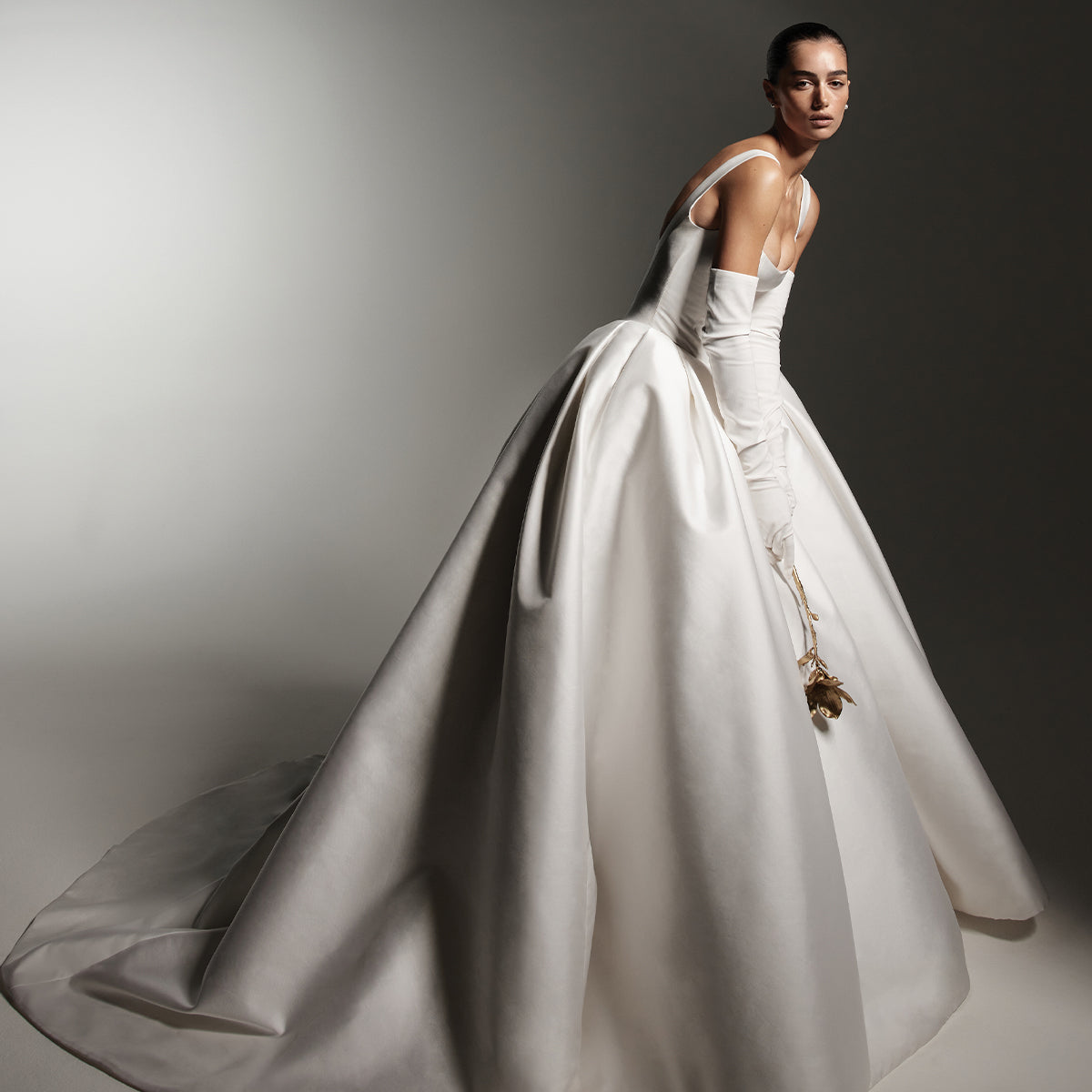 Model wearing Monique Lhuillier Spring 2026 Bridal white mikado Brentwood Scoop neck drop basque waist corset bodice ball
gown with white stretch crepe opera-length Chandler gloves, holding a gold flower