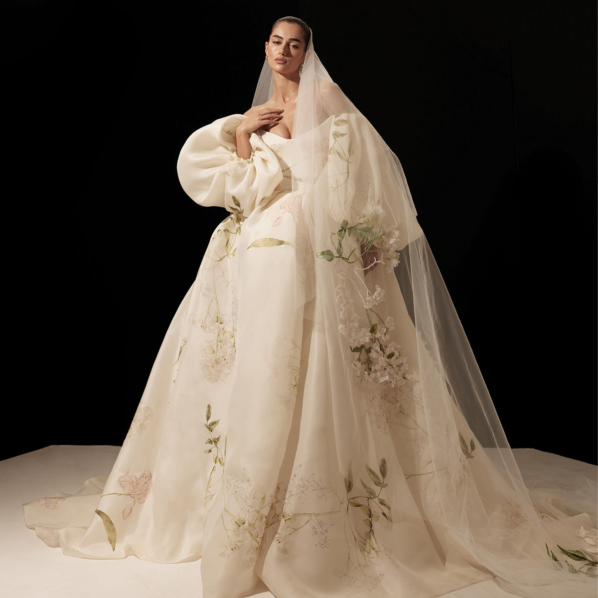 Model wearing Monique Lhuillier Spring 2026 Bridal ivory/multi floral printed gazar off-the-shoulder Layla ballgown with billow sleeves and voluminous skirt with slit, with floral embroidered white/multi two-tier long Layla veil