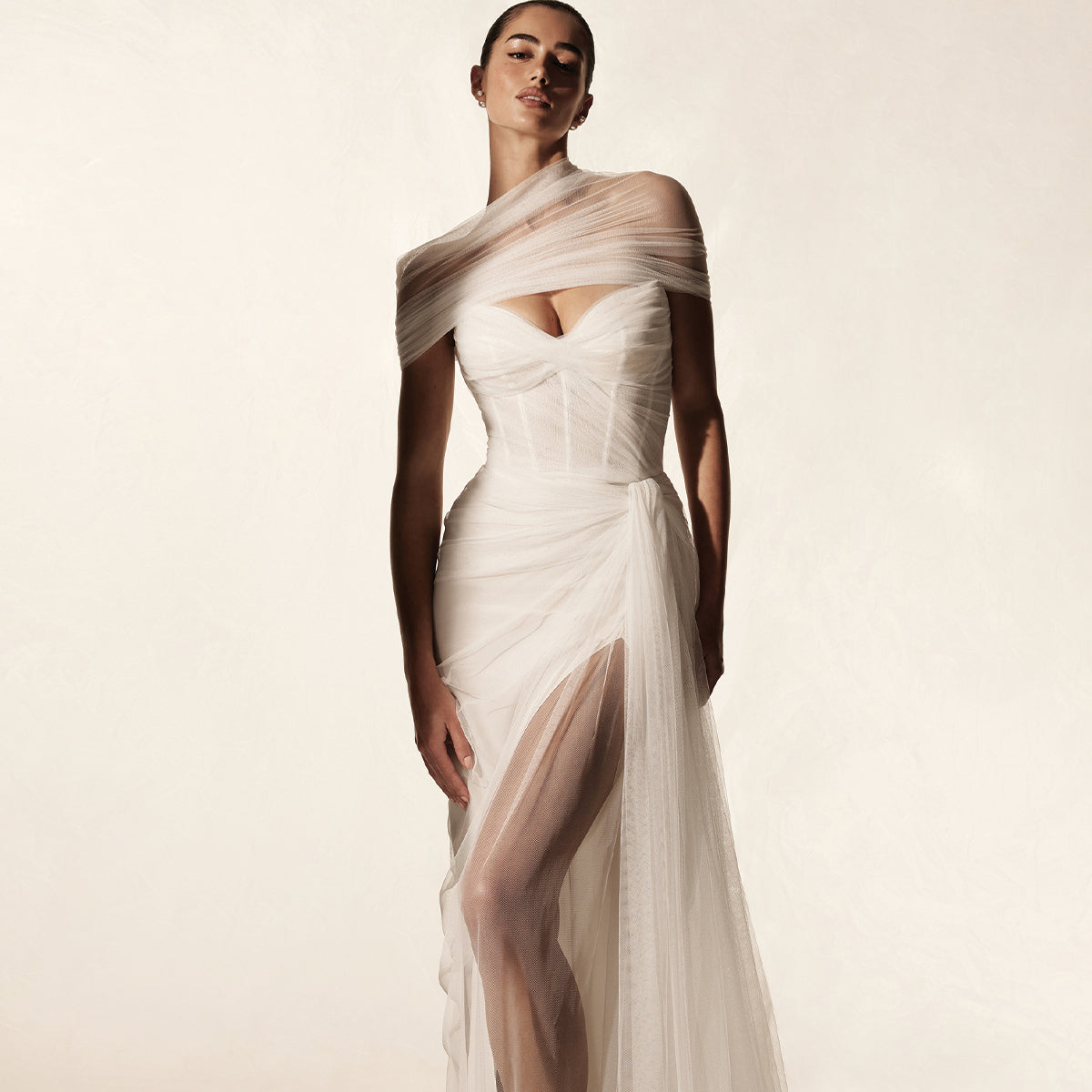 Model wearing Monique Lhuillier Spring 2026 Bridal white Spanish tulle strapless draped Sunset gown with sheer corset bodice and high skirt slit and matching Sunset wrap around shoulders