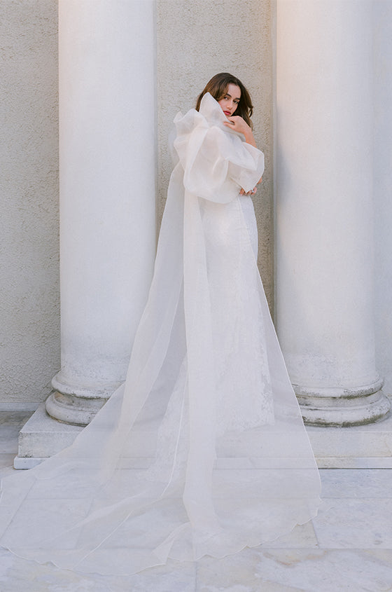 Woman wearing Monique Lhuillier Spring 2024 strapless white lace corseted Eden slip gown with puffed sleeve Barrington coat