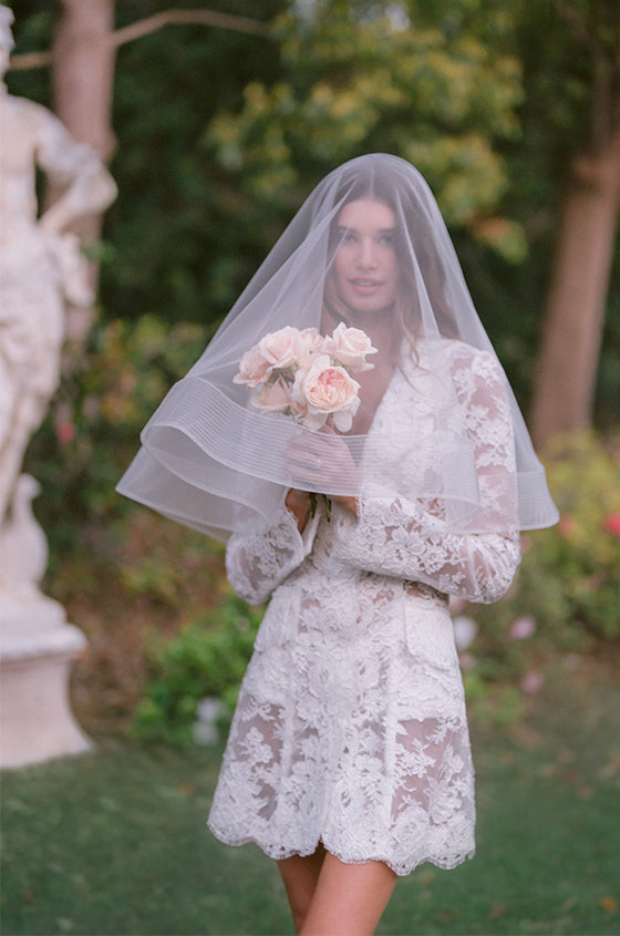 Woman wearing Monique Lhuillier Spring 2024 white lace long sleeve tailored A-line coat Claude cocktail dress with tulle veil over face