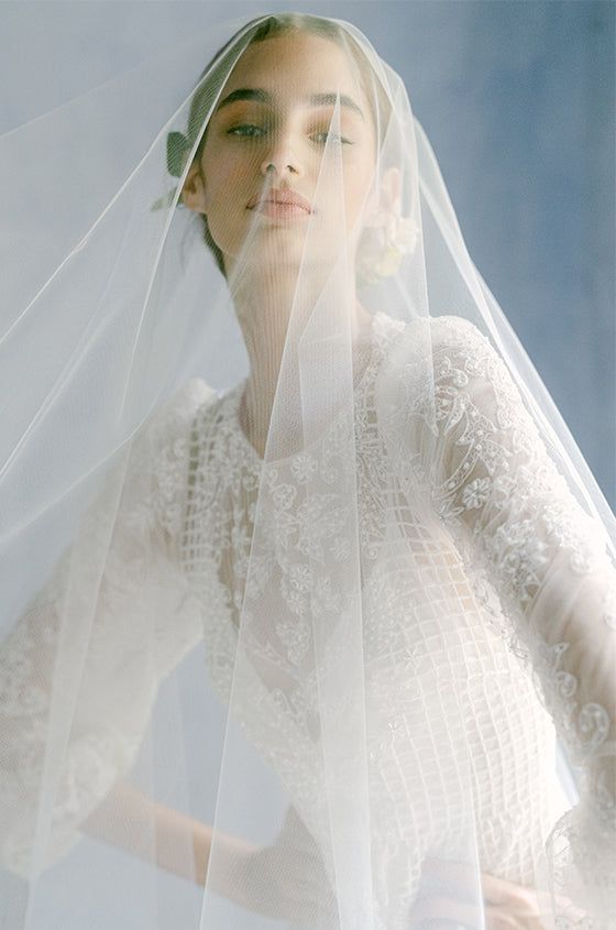 Woman wearing Monique Lhuillier Fall 2022 long sleeve white beaded sheath Emma gown with sheer veil over face
