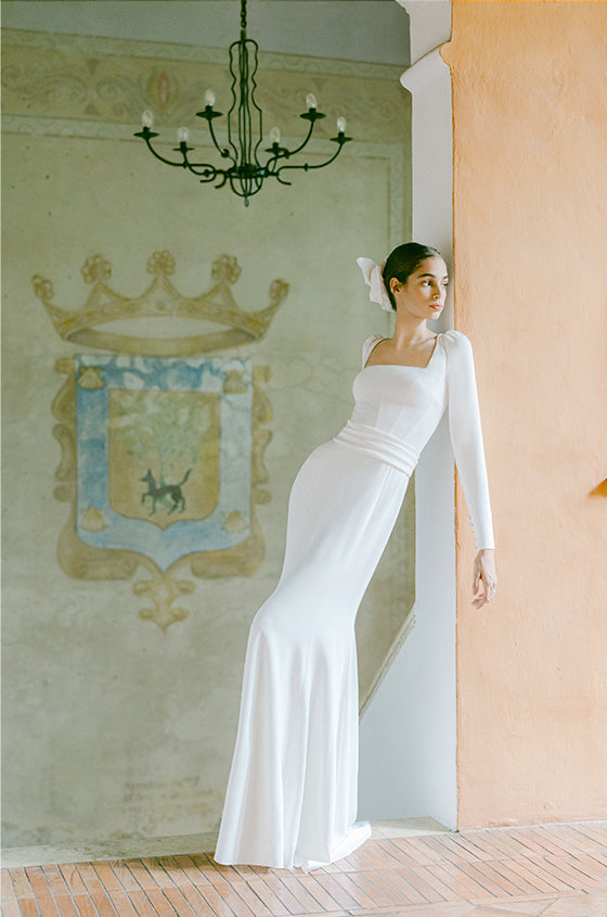 Woman wearing Monique Lhuillier Fall 2022 long sleeve white crepe sheath Phoebe gown with matching belt
