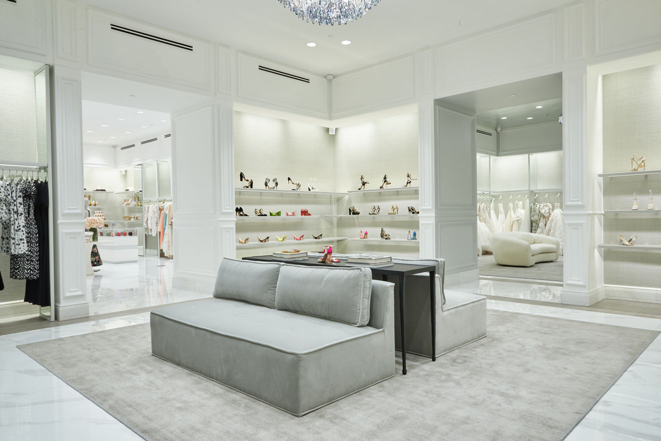 Interior of South Coast Plaza flagship store, featuring shoes, bridal gowns, and Ready to Wear with a grey velvet couch and carpet, white marble floor and crystal chandelier.