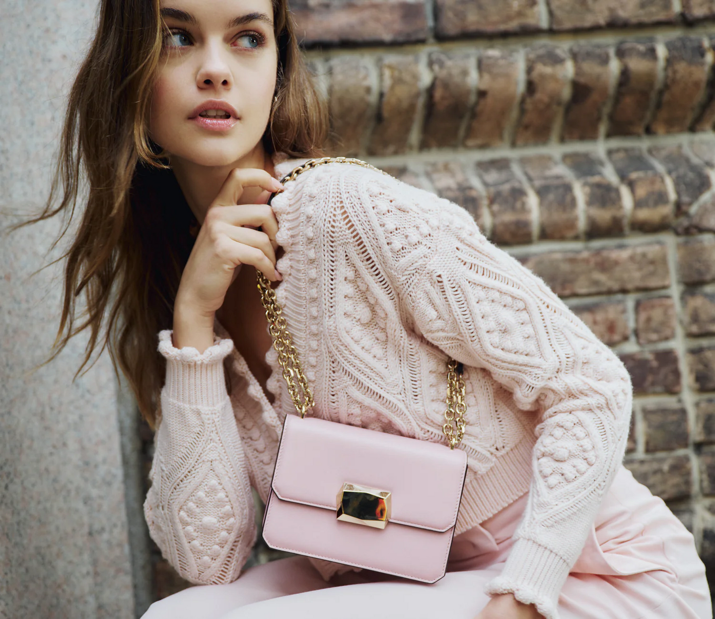 Model wearing blush Monique Lhuillier Fall 2024 Cable Knit Cardigan and trouser with blush Rhea mini shoulder bag with gold chain strap. 