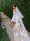 Woman wearing Monique Lhuillier Fall 2024 off-the-shoulder white long sleeve embroidered lace Kayla cocktail gown with tulle and lace veil