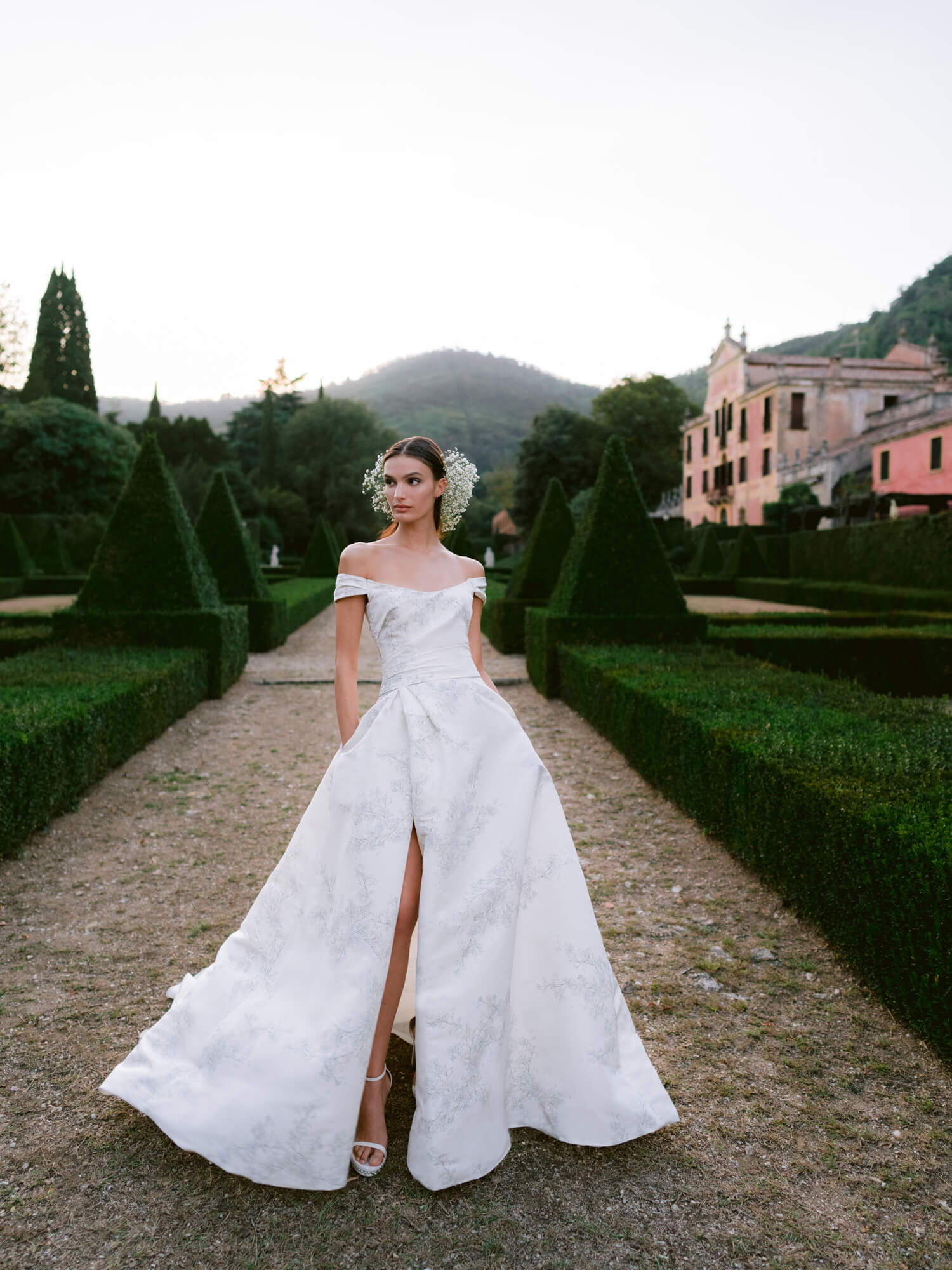 Woman wearing Monique Lhuillier Fall 2024 off-the-shoulder white printed faille Baby's Breath ballgown with slit