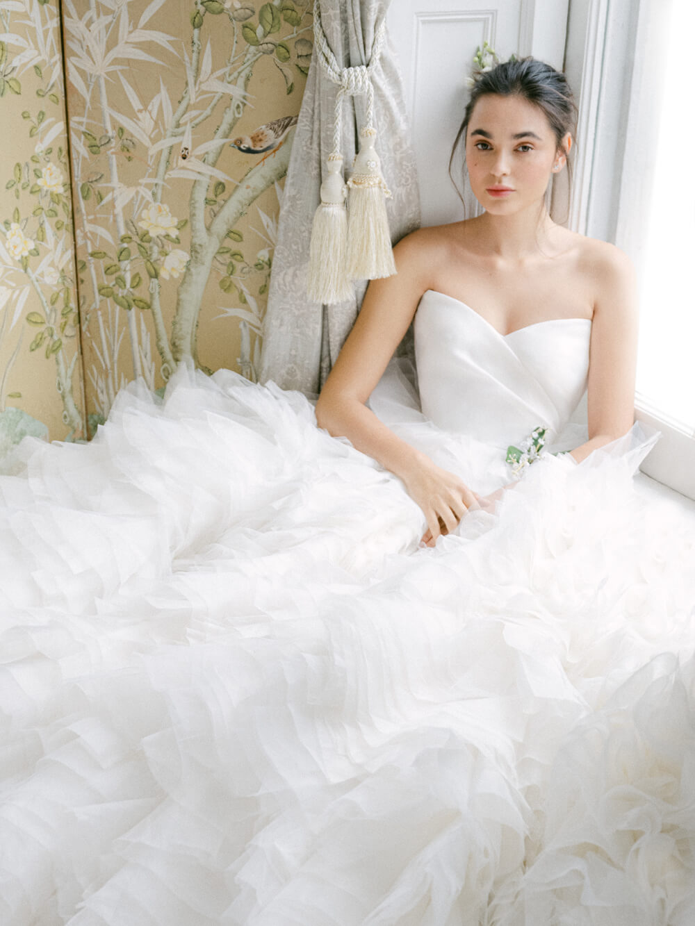 Model sitting in white strapless Daydream gown with organza layered ruffle skirt and lily of the valley brooch