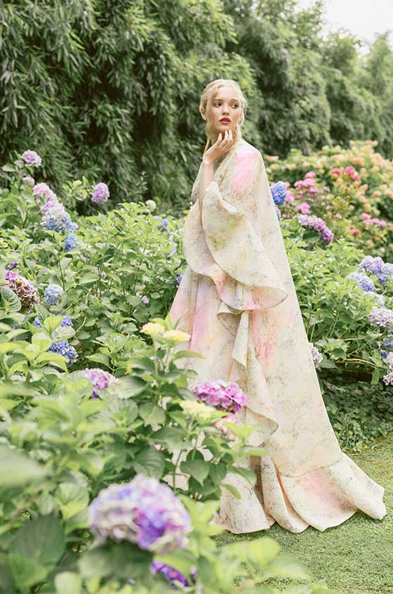 Woman wearing Monique Lhuillier Spring 2022 blush and green floral printed a-line gown and jacket with ruffle trim styles 22127 and 22128