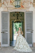 Woman wearing Monique Lhuillier Spring 2022 ivory and green lily of the valley printed halter neck Serena ballgown with long train