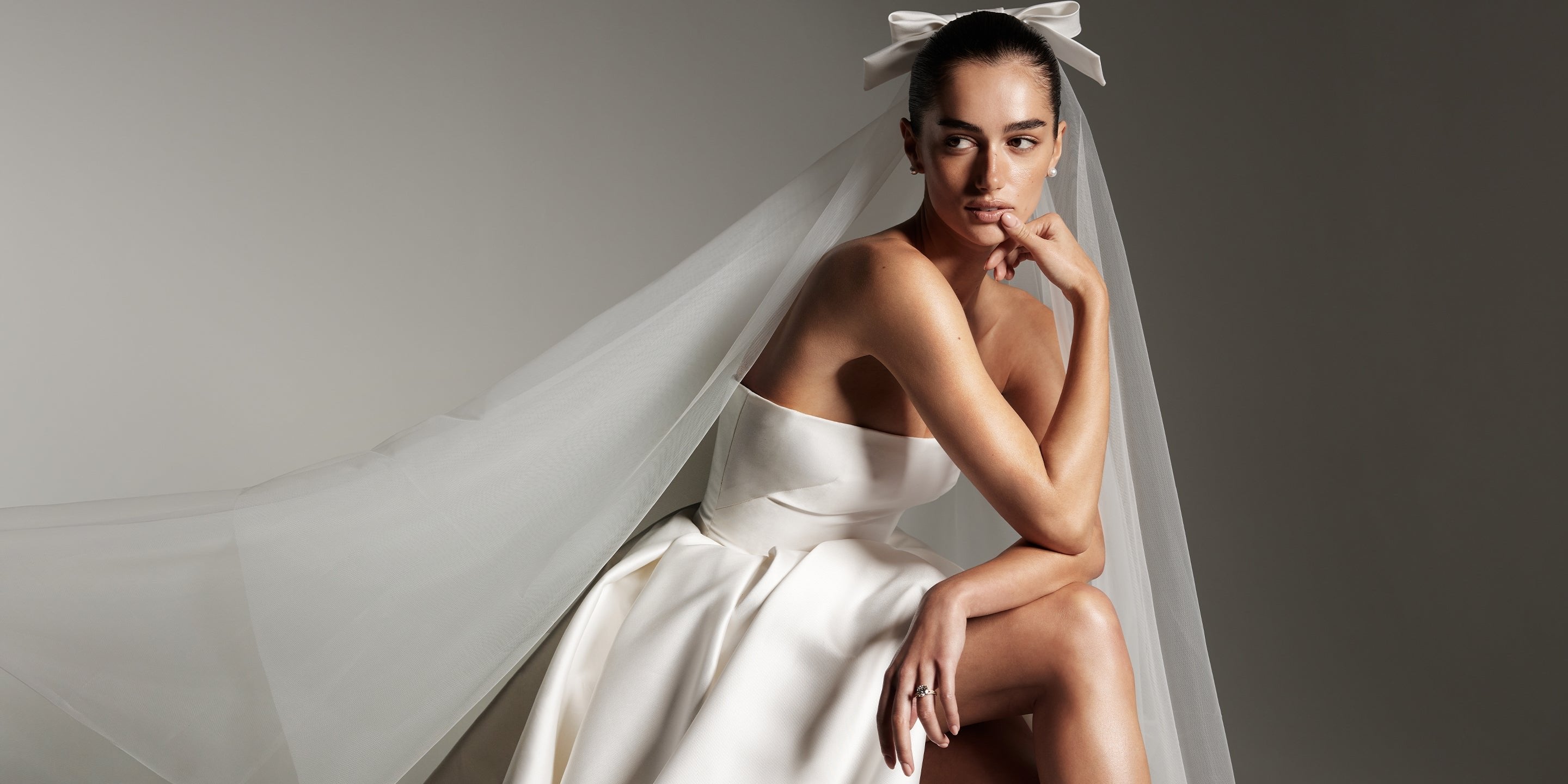 Model wearing Monique Lhuillier Spring 2026 Bridal white mikado Doheny strapless ball gown slit and two tier Doheny veil with bow at comb