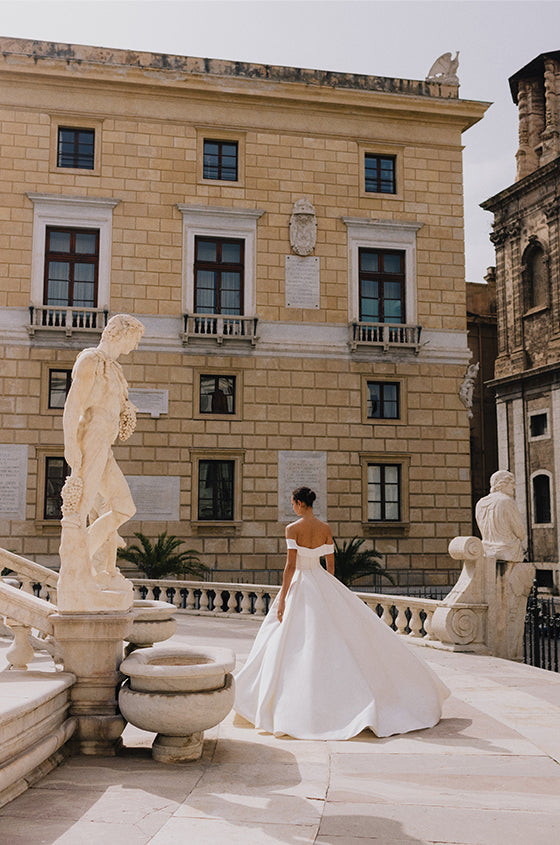 Woman wearing Monique Lhuillier Spring 2023 Bliss white off-the-shoulder mikado a-line BL23109 gown with side slit in skirt