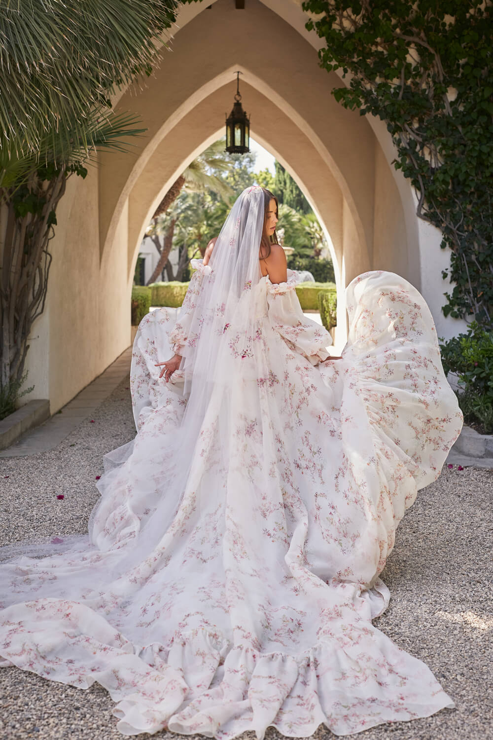 Woman walking in Monique Lhuillier Spring 2021 high-low, blush floral print strapless Valencia ballgown with long train