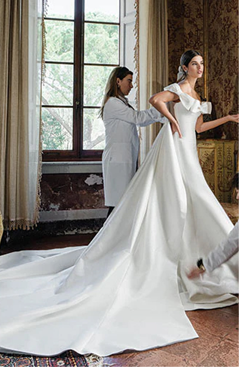 Monique Lhuillier Spring 2025 white mikado off-the-shoulder Mayfair trumpet gown with bow detail at neckline and train on model by window - lookbook.