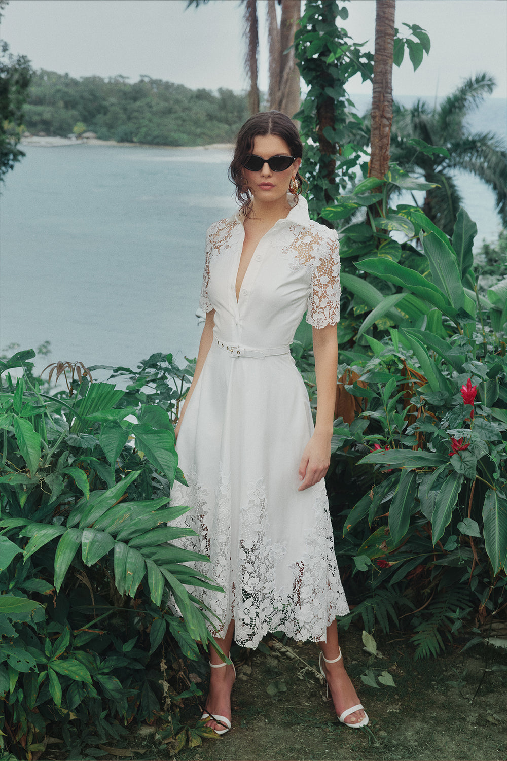 Monique Lhuillier Spring 2025 white cotton poplin dress with lace applique inserts, short sleeves, button down front bodice and belted waist - lookbook photo on model in grass.