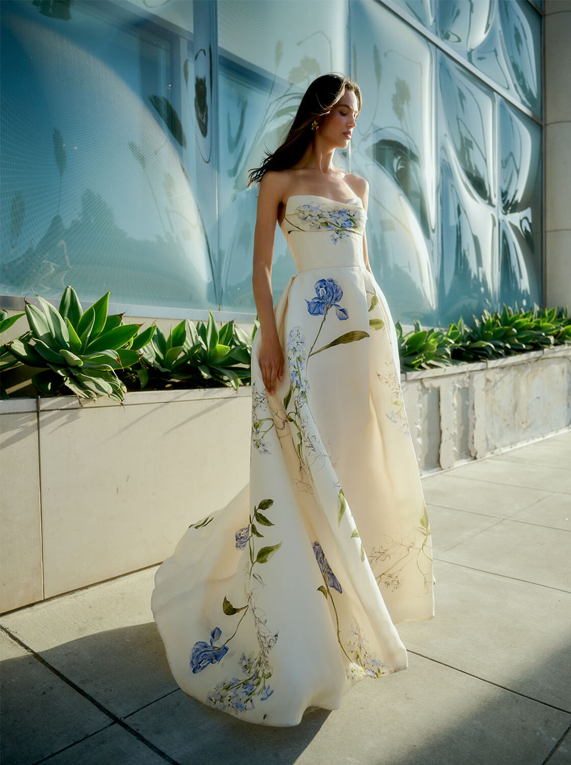 Monique Lhuillier Spring 2026 Ivory Multi Iris Bouquet Printed Gazar Draped bodice
ball gown with front skirt slit - model on sidewalk outside.