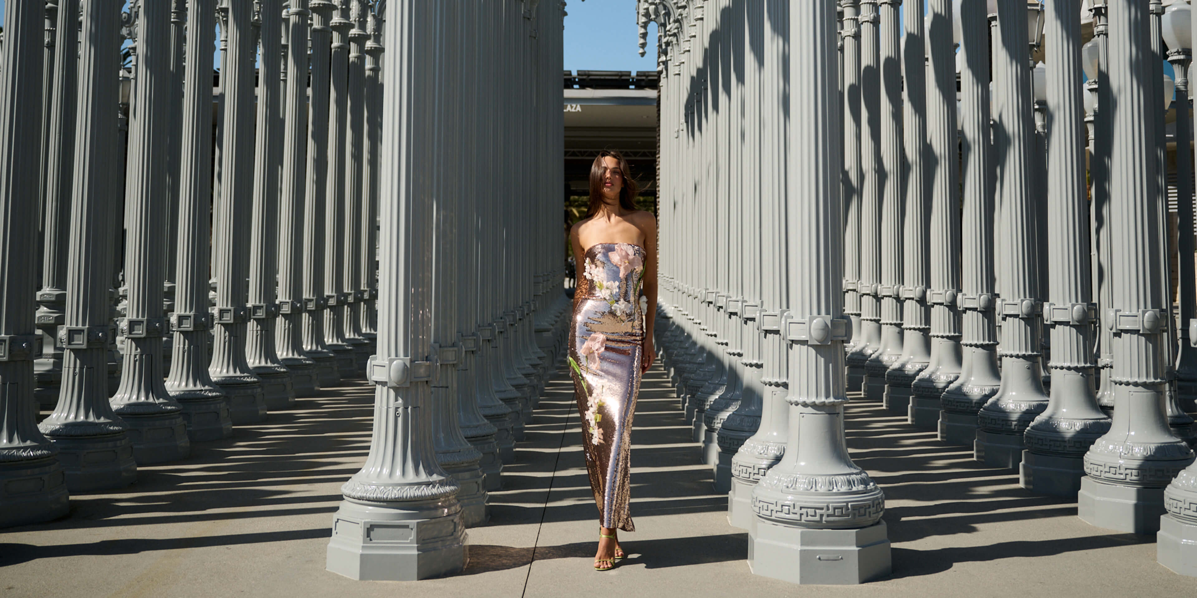Monique Lhuillier Spring 2026 Rose Gold Iris Bouquet Embroidered Sequin Strapless column gown with floral embroidery - model standing amongst lamp post light display.