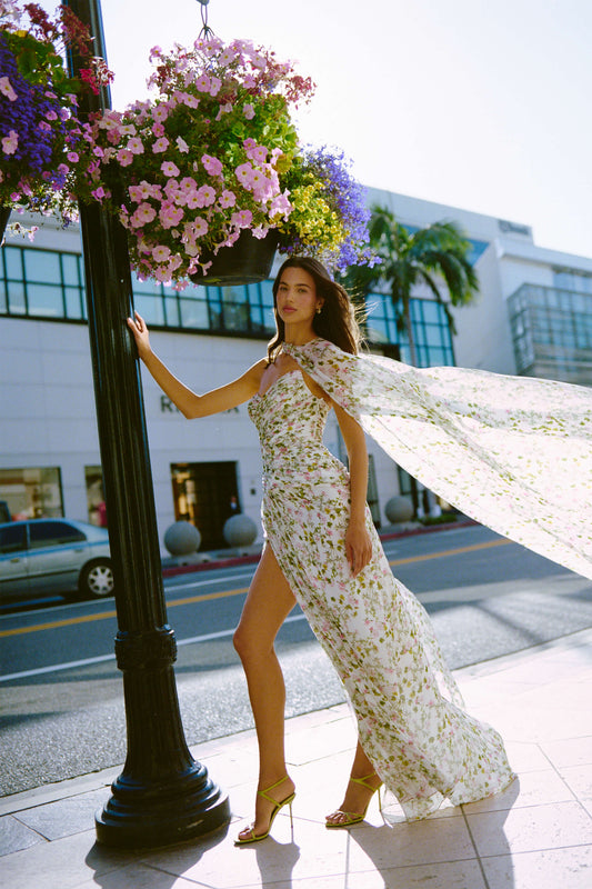 Monique Lhuillier Fall 2026 Silk White/Multi Ivy Bloom Printed Silk Chiffon Strapless asymmetric draped bodice gown with front slit - model on sidewalk outside. 