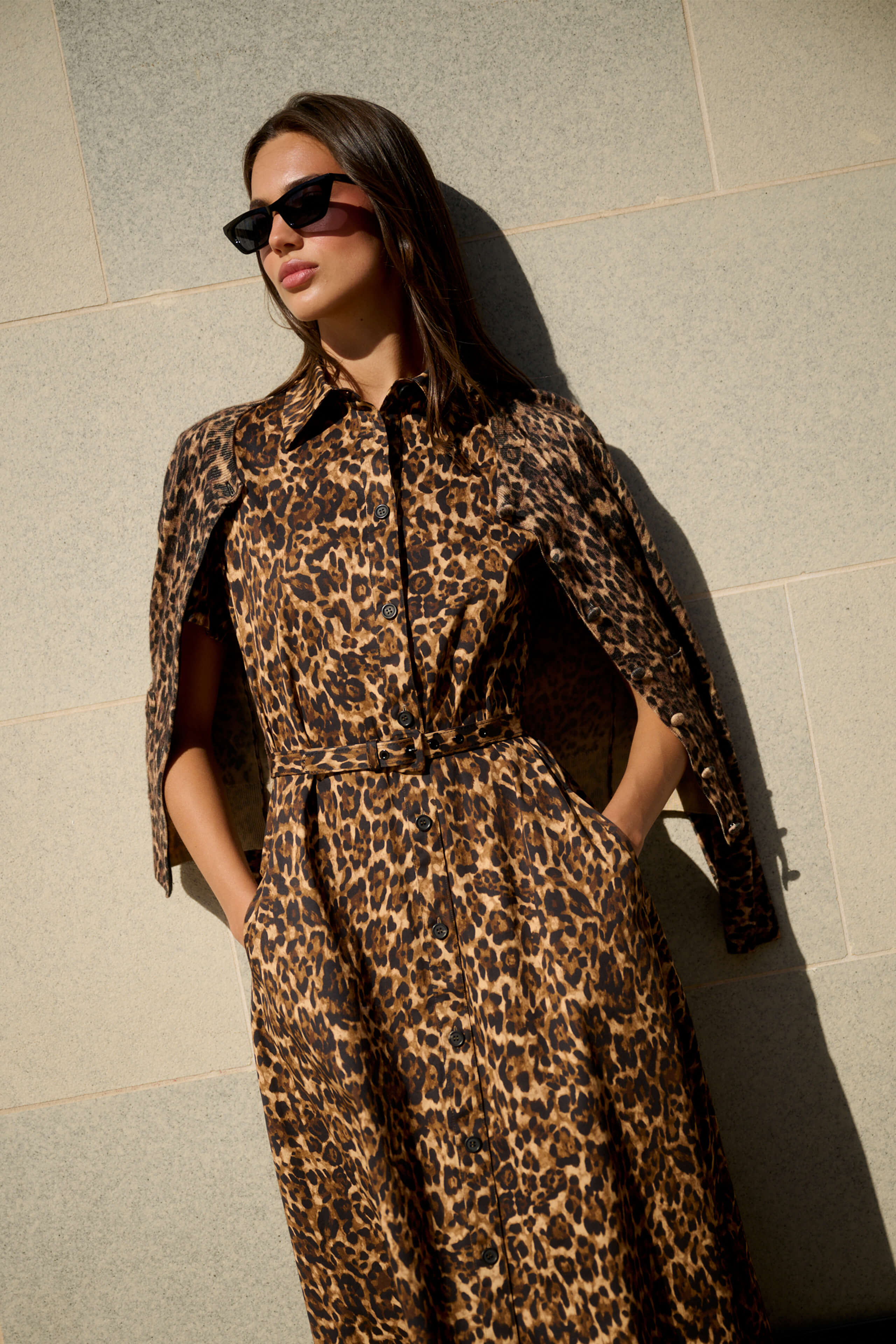 Monique Lhuillier Spring 2026 Leopard Printed Cotton Poplin short sleeve shirt
dress with belt and leopard cashmere cardigan - model leaning on wall outside.