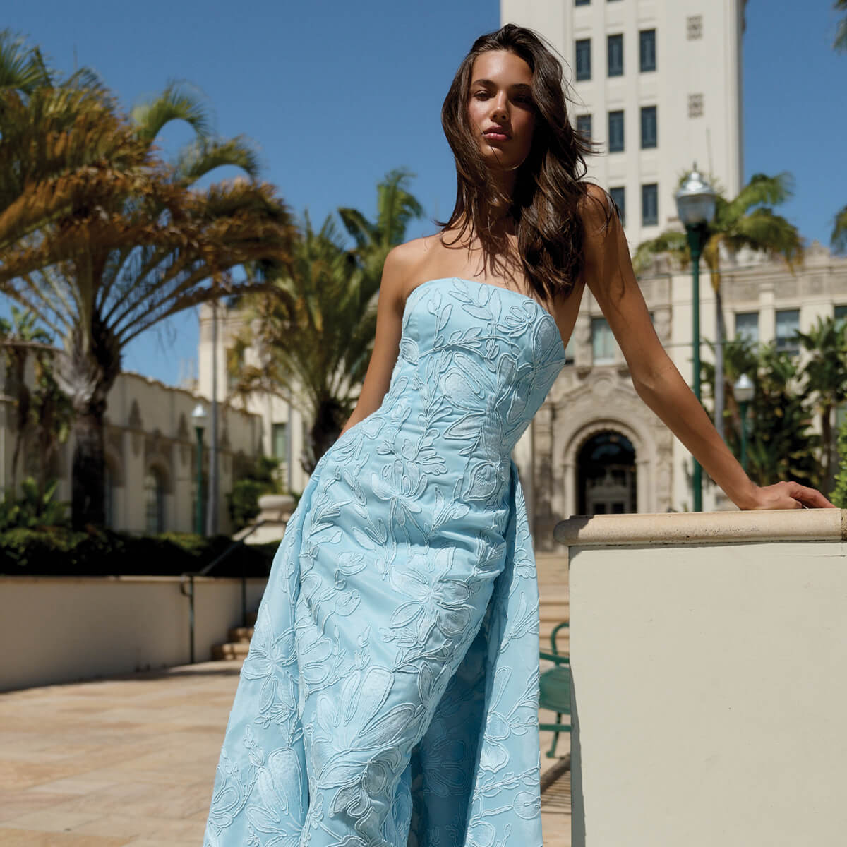 Monique Lhuillier Spring 2026 Plumeria Floral Corded Organza Strapless tea - length column gown with train - model leaning on ledge outside. 