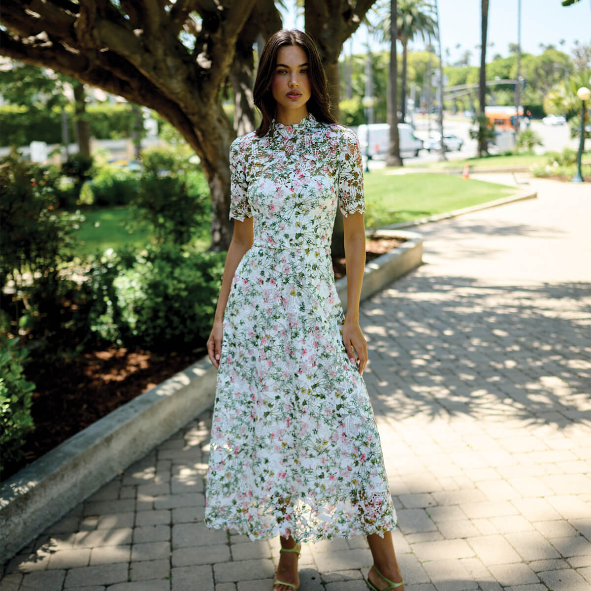 Monique Lhuillier Fall 2026 Silk White/Multi Ivy Bloom Printed Lace Short sleeve day dress with high neckline - model on stone walkway outside. 