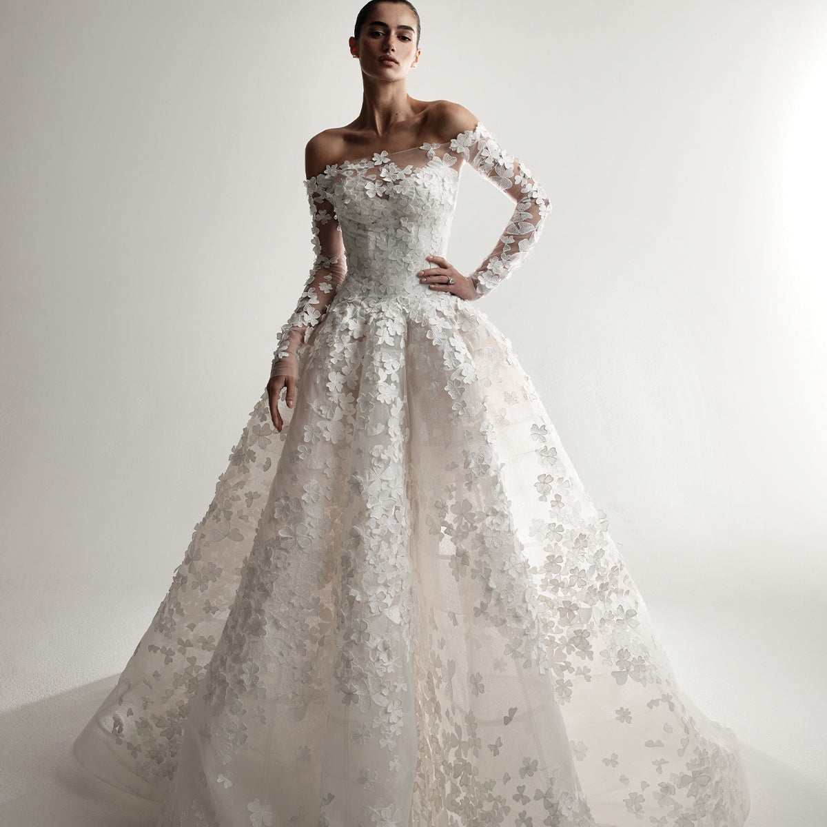 Model wearing Monique Lhuillier Spring 2026 Bridal white 3D embroidered tulle strapless elongated drop waist Clover ball gown with structured cone skirt and coordinating off-the-shoulder embroidered long sleeve Clover jacket 