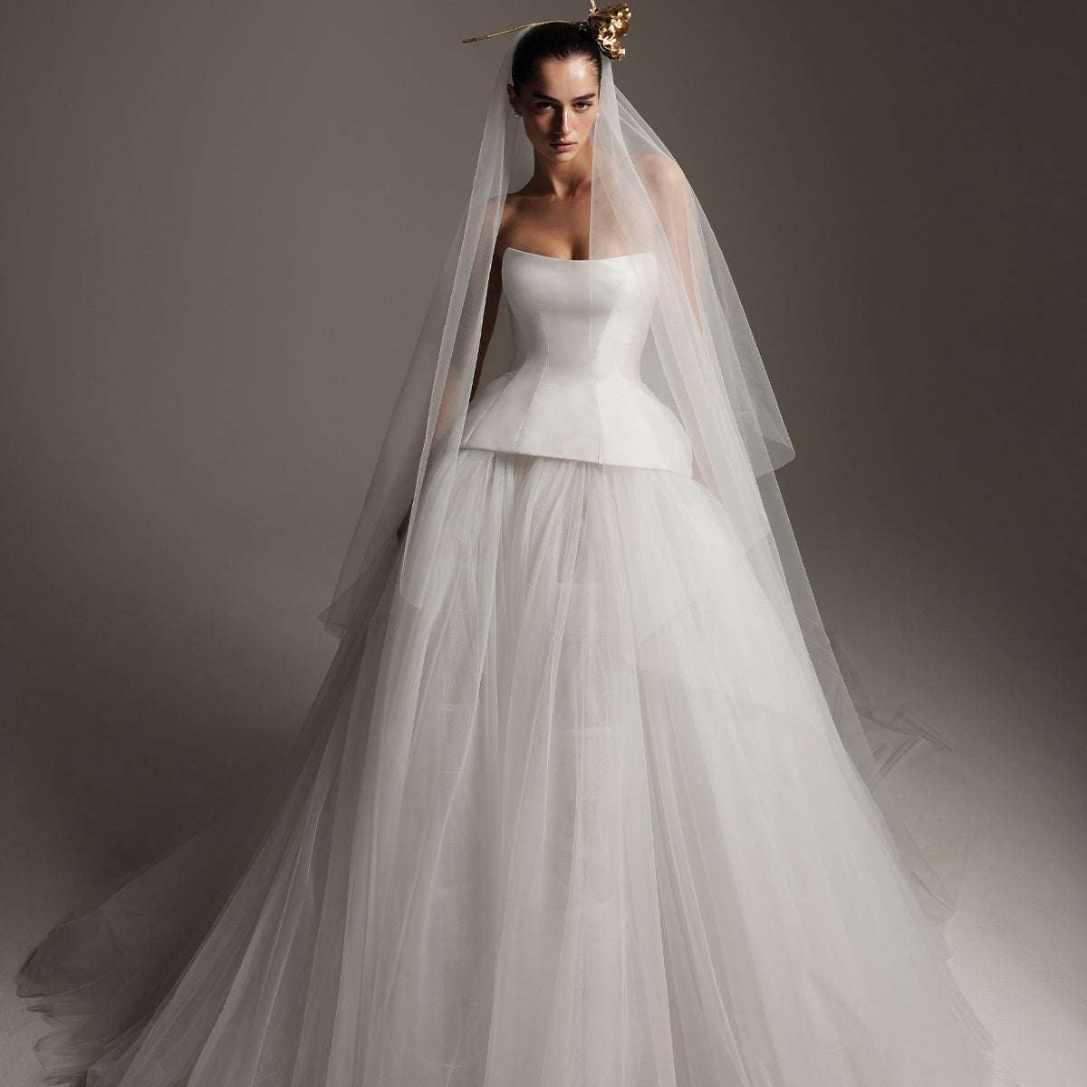 Model wearing Monique Lhuillier Spring 2026 Bridal Zuma white mikado and tulle strapless drop waist structured peplum corset with tulle ball gown skirt, with white tulle two-tier long veil and gold flower in hair 