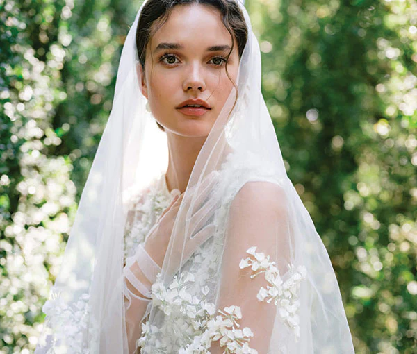Woman wearing Monique Lhuillier Fall 2024 white fitted sleeveless 3-d embroidered Gabrielle gown with matching veil and tulle gloves